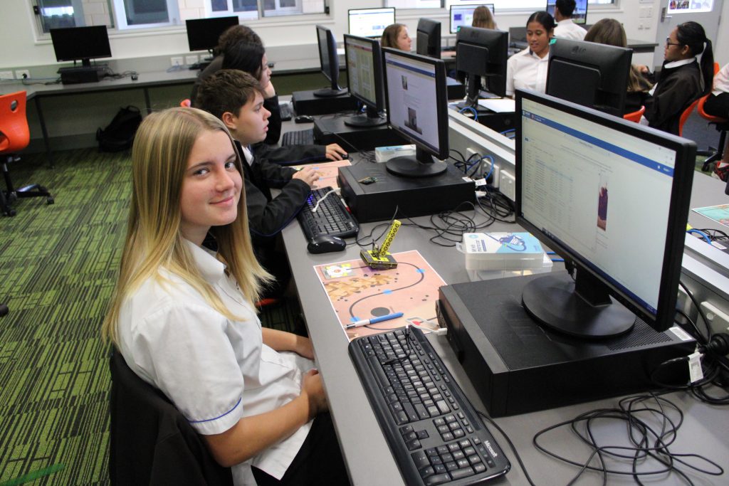 A CLC student in a white shirt with long blond hair sitting in front of a computer smiling with the microbit she is coding